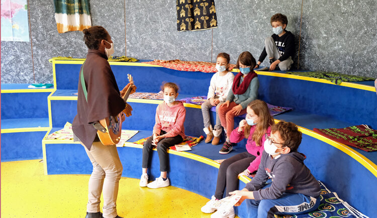 Une femme chante et joue ed la guitare avec un groupe d'enfant assis dans un peiti amphithéâtre en intérieur.