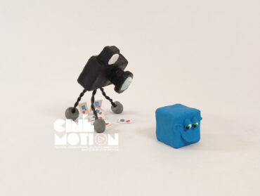 Un appareil photo sur pattes, réalisé en pâte à modeler, se penche pour regarder un cube bleu également en pâte à modeler.