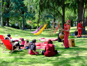 Une personne joue d'un instrument de musique devant des spectateurs assis sur des tapis et des chaises longues sur une pelouse. En arrière plan les arbres du Clos Fleuri.