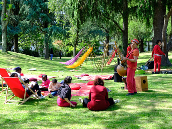 Une personne joue d'un instrument de musique devant des spectateurs assis sur des tapis et des chaises longues sur une pelouse. En arrière plan les arbres du Clos Fleuri.
