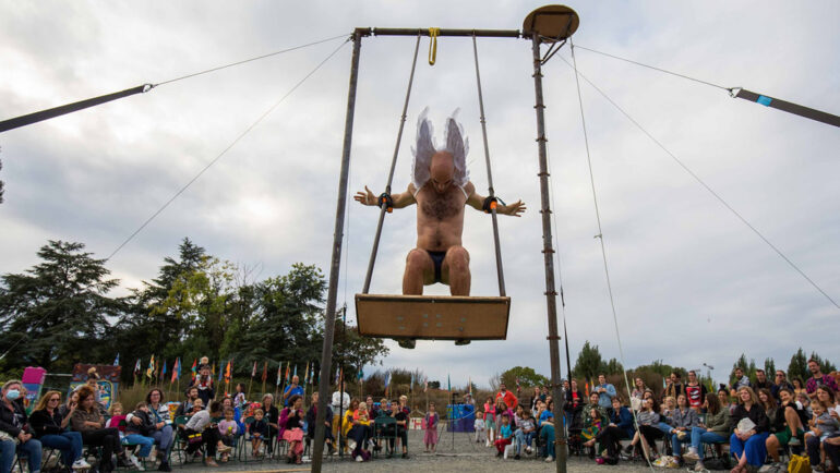 Un acrobate torse nu et portant des ailes d'ange se balance sur une balançoire géante au milieu des spectateurs.