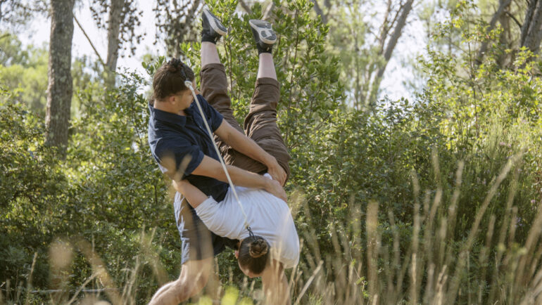 Deux personnes, liées par une corde tressée à leurs cheveux, sont accrochées l'une à l'autre. De shebes hautes au premier plan, une forêt à l'arrière plan.