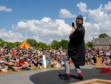 En premier plan on peut voir le clown qui regarde la caméra en faisant une grimace. En arrière-plan on peut voir le public assis par terre et attentif au spectacle.