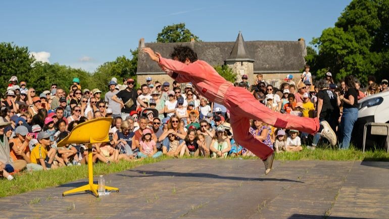 En premier plan nous pouvons voir le clown acrobatique portant une combinaison rouge. Il est en lévitation. En arrière-plan nous pouvons voir le public assis par terre et attentif au spectacle.