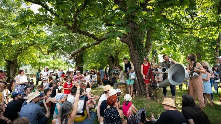 Au premier plan nous pouvons observer le public dos à la caméra entrain d’écouter et de regarder le spectacle. Au second plan nous pouvons voir le groupe Strohnofête entrain de performer.