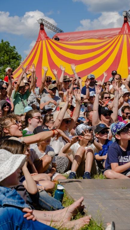 Au premier plan nous pouvons voir le public lever les mains et rire. Au second plan nous pouvons apercevoir le chapiteau du festival.