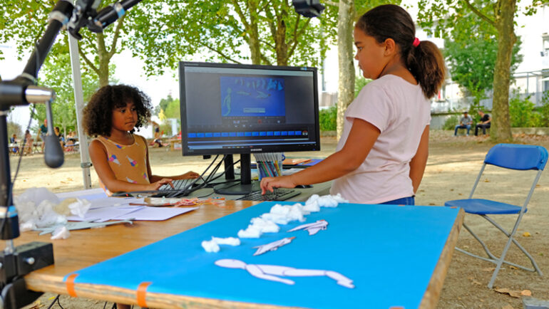 Deux enfants sont debout de part et d'autre d'un écran. Au premier plan une table avec des formes et personnages découpés et déposés sur une feuille.