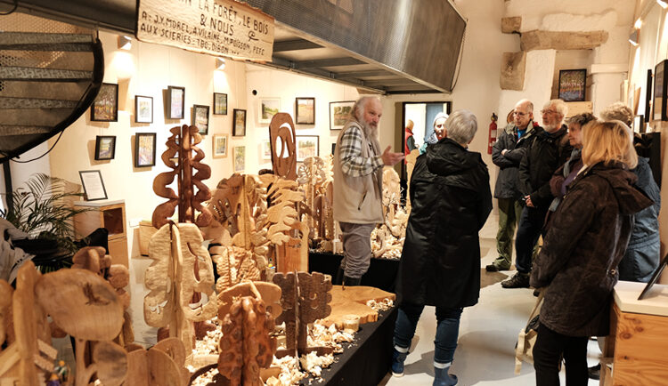 Un groupe de personnes écoute le sculpteur Paul Corbineau présenter son exposition sur le bois en cours à l'intérieur de la Longère.