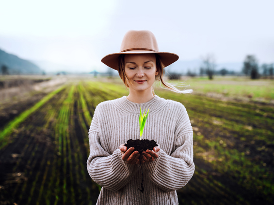 Debout dans un champ, une femme tient un motte de terre dans ses mains, dans laquelle pousse une plante.