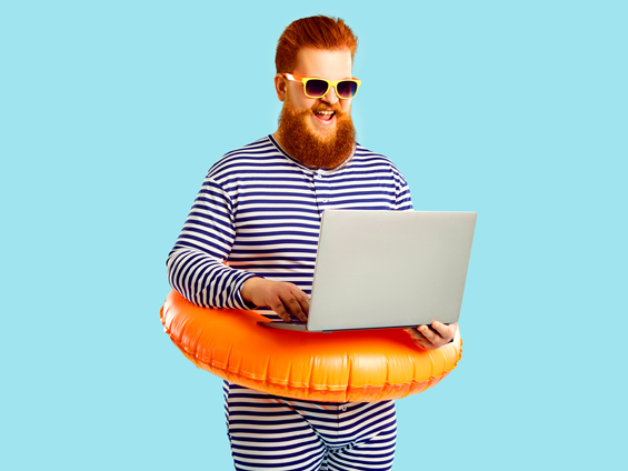 Un homme debout en barboteuse intégrale à imprimé marinière. Il a une bouée de plage autour du ventre et des lunettes de soleil et souris à son ordinateur portable qu'il porte à une main pendant qu'il pianote sur les touches.