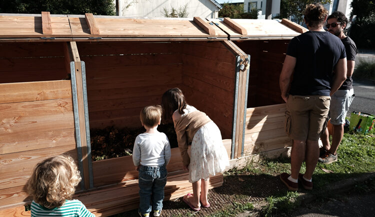 Deux enfants de dos observent le contenu d'un composteur collectif. Deux adultes discutent à côté.