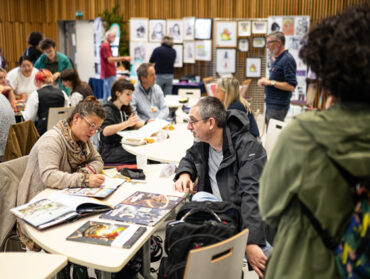 Plan large d'une salle occupée par des auteurs et autrices dédicaçant leurs ouvrages. En fond, on aperçois une exposition de dessins.