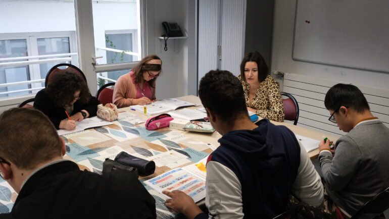 Dans une salle de classe, cinq adolescents et une institutrice autour d'une table, en cours de mathématiques.