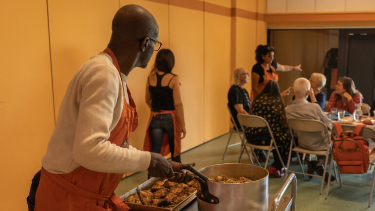 Au premier plan une personne amène un charriot chargé de nourriture vers un table ou des convives sont installés.