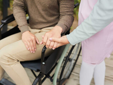 Une personne dans un fauteuil roulant donne la main à une personne debout dans un geste affectueux.