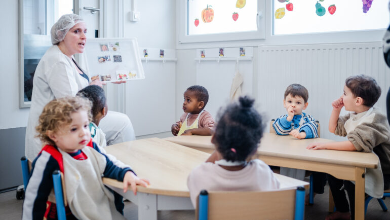 De très jeunes enfants sont autour d'une table. Avant le déjeuner, l'agent de restauration leur détaille le menuà l'aide d'images et mots épinglés sur un tableau.